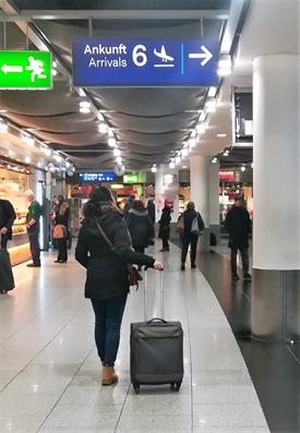 Eine junge Frau in dunkler Kleidung am Flughafen. Sie steht mit ihrem Koffer unter einem blauen Schild auf dem Ankunft steht und blickt nach oben auf das Schild.

